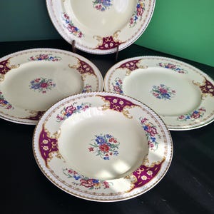 Può includere: Un set di quattro piatti in ceramica color crema con un bouquet floreale centrale di fiori rossi, blu e gialli. I bordi sono decorati con pannelli color magenta intenso, dettagli in filigrana dorata e motivi floreali con bordo oro.