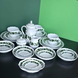 Puede incluir: Un juego de té de porcelana blanca con un estampado de hojas verdes. El juego incluye una tetera, una jarra de crema, un azucarero, tazas, platillos y platos de varios tamaños. Los bordes festoneados y los detalles verdes le dan un aire vintage.
