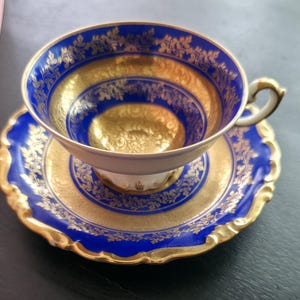 Op de afbeelding: Een porseleinen theekop en schotel set met een diep kobaltblauwe rand en ingewikkelde gouden bloemmotieven. De binnenkant van de kop en het midden van de schotel zijn afgewerkt met een glanzende goudkleurige glazuur en een geschulpte rand.