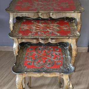 May include: A set of three nesting tables featuring a distressed gold finish on the curved legs and scalloped edges. Each tabletop has a rectangular surface with a vibrant red center inlay decorated with intricate, embossed gold scrollwork patterns.
