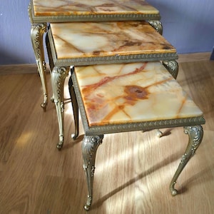 Op de afbeelding: Een set van drie rechthoekige bijzettafels met gepolijste amber- en crèmekleurige stenen bladen met natuurlijke aders. Elke tafel wordt ondersteund door vier sierlijke, gebogen, messingkleurige metalen poten met gedetailleerde randen.