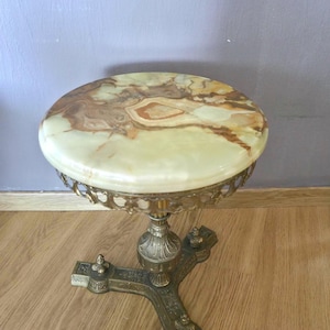 Op de afbeelding: Een ronde bijzettafel met een gepolijst, veelkleurig groen en bruin onyx stenen blad. De tafel wordt ondersteund door een sierlijke, goudkleurige metalen voet met drie decoratieve poten en gedetailleerd filigraanwerk rond de rand.