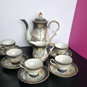 Op de afbeelding: Een vintage porseleinen theeservies met een theepot, melkkan, zes kopjes en schotels. De set heeft een zwart, grijs en goud kleurenschema met draken- en bloemmotieven. De theepot is ongeveer 20 cm hoog.