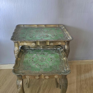Puede incluir: Un juego de dos mesas nido con tableros rectangulares de bordes festoneados y superficie verde con intrincados patrones de volutas en tono dorado. Las mesas tienen patas curvas y adornos dorados con pequeños detalles circulares.