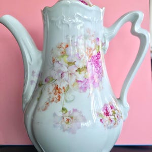 Op de afbeelding: Een witte porseleinen koffiepot met een delicaat bloemmotief in roze, oranje en groene tinten. De pot heeft een gebogen handvat, een lange tuit en een bijpassend deksel met een decoratieve knop, afgewerkt met subtiele reliëfranden.