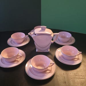 Peut inclure: Un service à thé composé d'une théière centrale avec un couvercle géométrique et quatre tasses assorties avec soucoupes. Chaque pièce est rose clair avec un fin liseré argenté sur les bords. La théière présente une forme angulaire distincte.