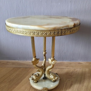 May include: A round side table featuring a polished cream and tan marble top with a decorative gold-toned metal rim. The base consists of three vertical fluted columns supported by three brass dolphin figures resting on a circular marble pedestal base.