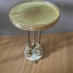 May include: A round side table featuring a pale green stone top with a gold-toned metal lattice gallery edge. The base consists of three vertical metal columns supported by three brass dolphin figures resting on a circular white marble base.