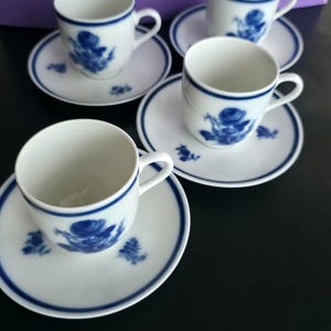 Puede incluir: Un juego de cuatro tazas de espresso de porcelana blanca con sus platos a juego, cada una con un patrón de rosas azules y un borde fino azul. Las tazas tienen una pequeña asa lateral y los platos muestran un motivo floral azul a juego.