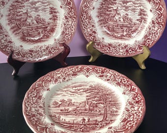 May include: Three round ceramic plates featuring a deep red transferware pattern on a white background. Each plate displays a detailed pastoral landscape scene in the center, surrounded by a wide border of intricate floral and lattice motifs.
