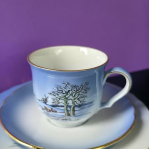 Puede incluir: Un juego de taza y platillo de porcelana con acabado en degradado azul claro y borde dorado. La taza muestra una ilustración de paisaje invernal con árboles desnudos y un pequeño molino. El platillo tiene un borde azul a juego y filo dorado.