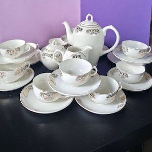 Op de afbeelding: Een wit porseleinen theeservies bestaande uit een theepot, suikerpot, melkkan en meerdere theekopjes met bijpassende schoteltjes. Elk stuk is versierd met een delicaat goudkleurig bloemmotief langs de randen en zijkanten, afgewerkt met goud.