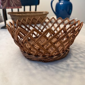 May include: A round, brown woven basket featuring a decorative crisscross lattice pattern along the sides and a solid, circular base. The rim of the basket has a distinct zigzag edge, and the material appears to be a natural, flexible reed or wicker.