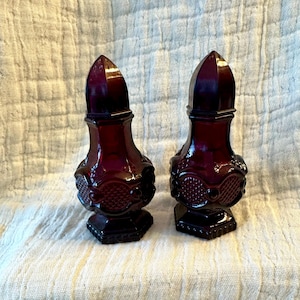 Op de afbeelding: Twee antieke, donkerrode glazen parfumflesjes. Elk flesje heeft een facetgeslepen, puntige stopper en een decoratief, getextureerd lichaam. De flesjes staan op een vierkante basis. Het glas heeft een glanzende afwerking.