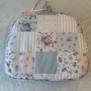 May include: A patchwork chair cushion with a variety of floral, striped, and polka dot patterns in pastel colors. The cushion has a rounded shape and a light blue piping around the edge. It features a handle made of fabric.