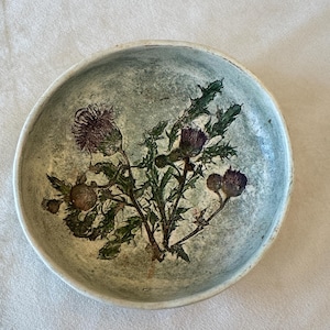 May include: A round, ceramic bowl with a light green and gray mottled finish. The bowl's interior features a detailed botanical print of a thistle plant with purple flowers and green leaves. The rim is a lighter shade of green.