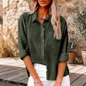 Peut inclure: Une chemise boutonnée vert olive à manches longues avec un col et une poche poitrine. La chemise est portée avec un pantalon blanc. Les manches sont retroussées. En arrière-plan, un mur en pierre et des plantes en pot.