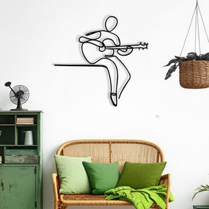 May include: Black metal wall art depicting a musician playing a guitar. The minimalist design shows a figure seated, with the guitar in their lap. The art piece is mounted on a white wall, above a wicker bench with green pillows.