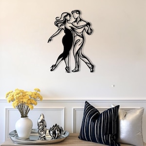 Puede incluir: Una pieza de arte mural de metal negro que representa a una pareja bailando. La obra muestra a un hombre y una mujer abrazados, con la mujer con un vestido largo y tacones. La pieza está montada en una pared blanca, creando un contraste llamativo.