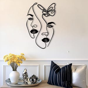 May include: Black metal wall art featuring two stylized faces and a butterfly. The artwork is a continuous line design, with bold black lines defining the facial features. The piece is mounted on a white wall, creating a striking contrast.