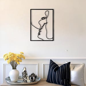 May include: Black metal wall art depicting two faces in a continuous line design. The artwork is framed in a rectangular black border and mounted on a white wall. The image also shows a table with decorative items and pillows.