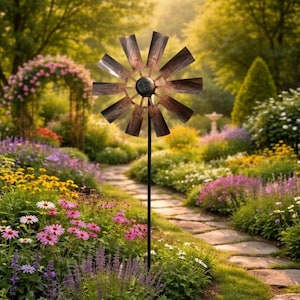 Puede incluir: Una estaca de jardín de metal rústico con forma de molino de viento y acabado marrón desgastado. El molino de viento presenta un diseño circular con múltiples aspas y un cubo central negro. La estaca está colocada en un jardín exuberante con un sendero de piedra y flores coloridas.