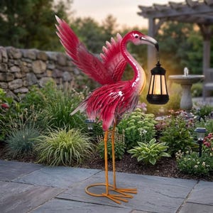 Puede incluir: Una estatua de jardín de metal con forma de flamenco, en tonos rosa y rojo, con las alas extendidas. Sostiene una linterna negra con luz cálida. El flamenco tiene largas patas naranjas y está en un jardín.
