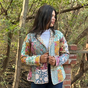 May include: A patchwork jacket with floral and geometric patterns in shades of blue, pink, yellow, and green. The jacket has a collared neckline and long sleeves. The jacket is worn over a white top and blue jeans.