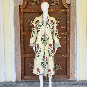 Puede incluir: Una bata larga de color crema con un estampado floral de rosas rojas y hojas verdes. La bata tiene mangas largas y un detalle de puño a rayas. La bata se exhibe en un maniquí frente a una puerta de madera.