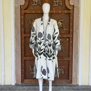 Könnte beinhalten: Ein langer, offener Mantel mit floralem Muster. Der Mantel ist weiß mit grauen und schwarzen Blumenmustern und grünen Akzenten an den Ärmeln. Der Mantel wird auf einer Schaufensterpuppe vor einer Holztür präsentiert.