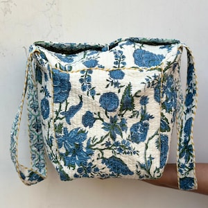 May include: A quilted tote bag with a floral pattern in shades of blue and green on a cream background. The bag has a rectangular shape with a top opening and long straps. The straps are trimmed with a gold-colored material.