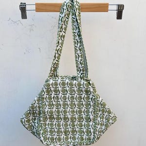 May include: A white tote bag with a green floral pattern. The bag has two handles and is hanging from a wooden hanger. The bag's design features a repeating pattern of green leaves and flowers.