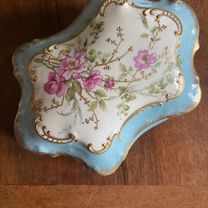 May include: A rectangular porcelain trinket box with a light blue border and a white center panel. The lid features a hand-painted floral design with pink roses, green leaves, and gold filigree accents along the scalloped edges of the box.