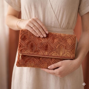 May include: A rust-coloured velvet fold-over clutch bag featuring a raised, geometric, fan-like pattern across the surface. The rectangular bag includes a horizontal metal zip closure along the fold line with a small, matching metal zip pull.
