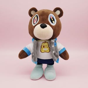 May include: A plush brown bear wearing a white shirt with a graphic, a grey jacket, blue trousers, and light blue shoes. The bear has large eyes with colourful details and is standing against a pink background. A collectible toy.