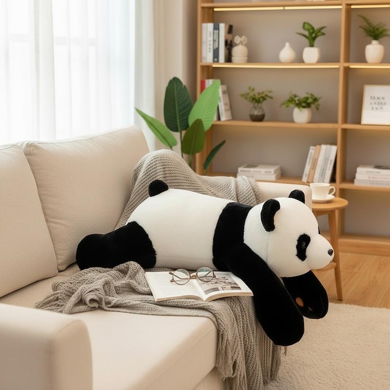 Puede incluir: Un gran peluche de oso panda tumbado en un sof&aacute;. El panda es blanco con manchas negras en las extremidades, las orejas y alrededor de los ojos. Descansa sobre una manta beige con un libro abierto y gafas.