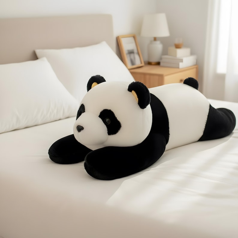 Puede incluir: Un peluche de oso panda en posici&oacute;n horizontal, con un dise&ntilde;o en blanco y negro. El panda tiene patas, orejas y parches oculares negros, con un cuerpo blanco. El juguete est&aacute; sobre una cama blanca.