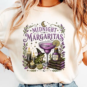 May include: A cream-colored t-shirt featuring a graphic print with the text "MIDNIGHT MARGARITAS". The design includes a purple margarita, a frog wearing a witch hat, three lit candles, a bottle labeled "LIQUID MISCHIEF", and stacked books.