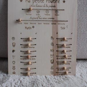 Peut inclure: Un tableau de routine quotidienne en bois clair avec des textes gravés en français, des icônes et des perles en bois coulissantes sur des fils métalliques. Le tableau comprend des sections pour l'humeur, les jours et les tâches.