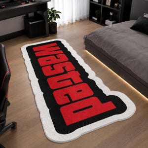 May include: A rectangular area rug with rounded corners featuring a white border, a thick black inner border, and the word WASTED printed in large, bold red capital letters. The rug has a soft, textured surface and a distinct, modern graphic design.