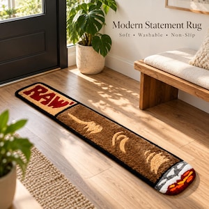 Peut inclure: Un long tapis rectangulaire en forme de cigarette, présentant le texte "RAW" en rouge sur une section filtre beige, un corps marron avec des accents beiges, et un bout incandescent blanc, orange et rouge, le tout bordé d'un liseré noir.
