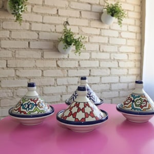 Puede incluir: Una colección de ollas de cerámica tagine con tapas cónicas, con coloridos patrones geométricos en rojo, azul, verde y blanco. Las ollas están sobre una mesa rosa contra una pared de ladrillos blancos, con plantas colgantes en el fondo.