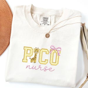 Puede incluir: Camiseta color crema con el texto bordado "PICO nurse" en cuadros amarillos y escritura rosa. Un dibujo animado de jirafa y un lazo rosa acent&uacute;an el dise&ntilde;o. Un collar dorado con un colgante est&aacute; drapeado sobre la camisa.