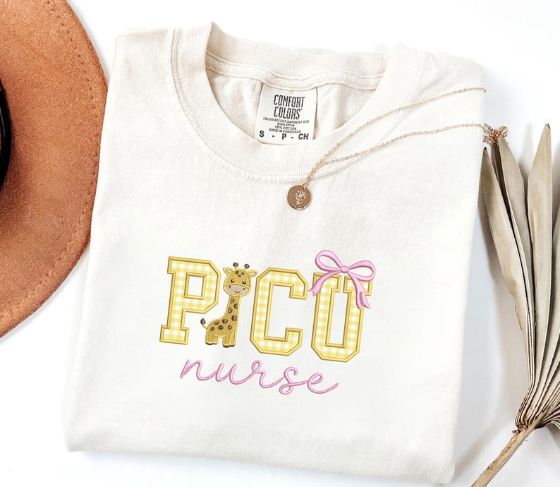 Puede incluir: Camiseta color crema con el texto bordado "PICO nurse" en cuadros amarillos y escritura rosa. Un dibujo animado de jirafa y un lazo rosa acent&uacute;an el dise&ntilde;o. Un collar dorado con un colgante est&aacute; drapeado sobre la camisa.