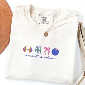 Puede incluir: Camiseta color crema con un gr&aacute;fico de una mancuerna, andador, lazo y bal&oacute;n de ejercicio. El texto "movement is medicine" est&aacute; debajo. Un collar dorado con un colgante est&aacute; sobre la camiseta.