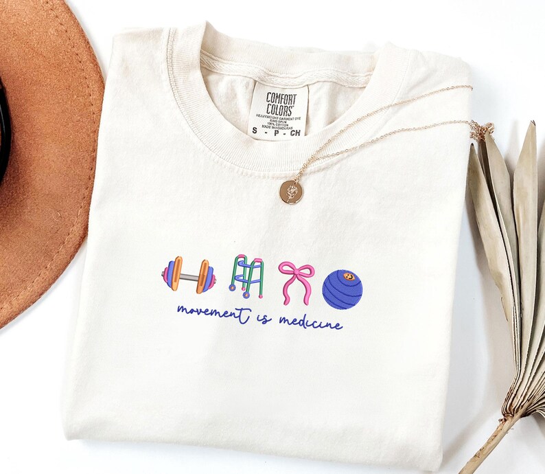 Puede incluir: Camiseta color crema con un gr&aacute;fico de una mancuerna, andador, lazo y bal&oacute;n de ejercicio. El texto "movement is medicine" est&aacute; debajo. Un collar dorado con un colgante est&aacute; sobre la camiseta.