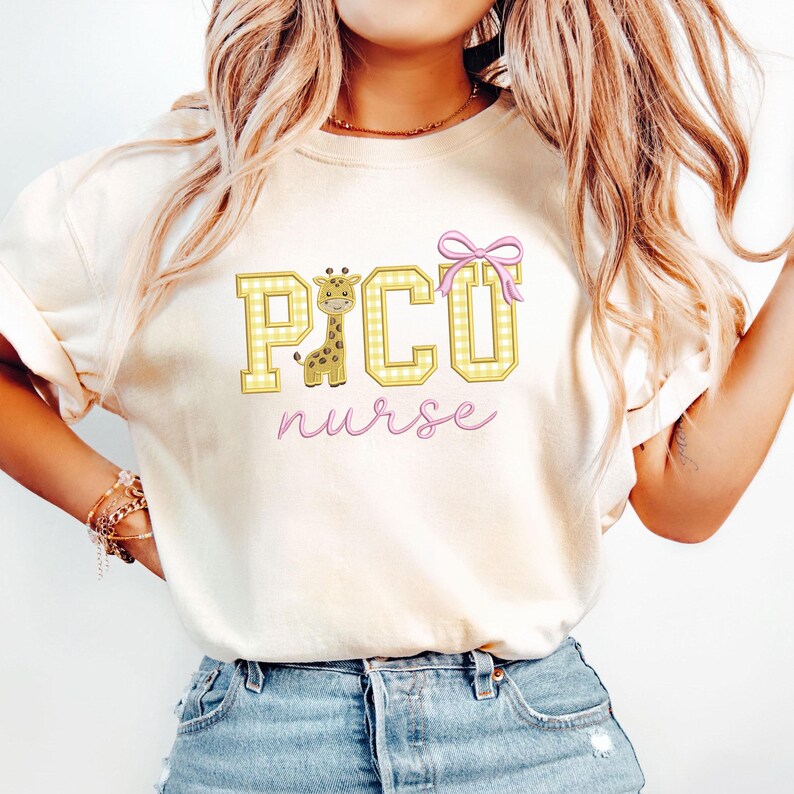 Puede incluir: Camiseta color crema con un dise&ntilde;o gr&aacute;fico. El dise&ntilde;o presenta la palabra "PICO" en letras de cuadros amarillos, una jirafa, un lazo rosa y la palabra "nurse" en cursiva. La camiseta tiene mangas cortas.