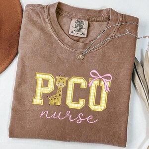 Puede incluir: Camiseta Comfort Colors marr&oacute;n con un gr&aacute;fico de jirafa y el texto "PICO nurse" en amarillo y rosa. La jirafa est&aacute; dentro de la letra "C". Un collar plateado con un peque&ntilde;o colgante est&aacute; sobre la camiseta.