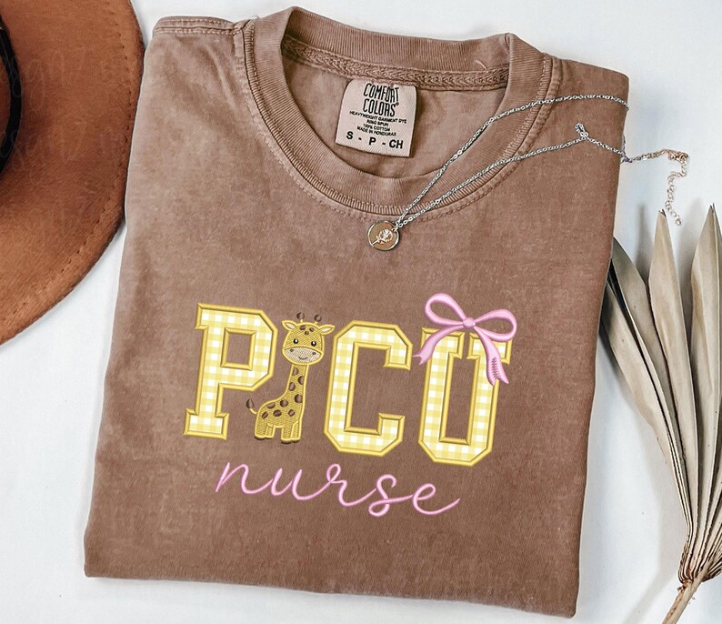 Puede incluir: Camiseta Comfort Colors marr&oacute;n con un gr&aacute;fico de jirafa y el texto "PICO nurse" en amarillo y rosa. La jirafa est&aacute; dentro de la letra "C". Un collar plateado con un peque&ntilde;o colgante est&aacute; sobre la camiseta.