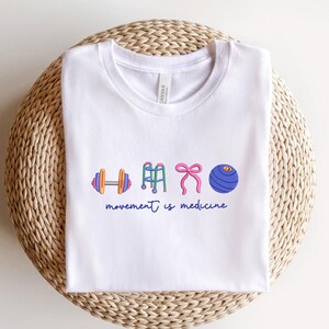 Puede incluir: Camiseta blanca con cuello redondo con ilustraciones coloridas de equipos de ejercicio: una mancuerna, un andador, un lazo rosa y una pelota de ejercicios. El texto "movement is medicine" est&aacute; escrito en azul debajo.